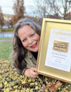En leende Eva Marie Svensson ligger på mage bland fallna höstlöv och tittar fram bakom pristavlan med guldkassetten.